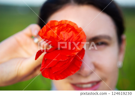 A smiling woman playfully covers her eye with a vibrant red poppy flower, enjoying a sunny day outdoors 124619230