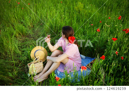 A woman enjoys a picnic in a vibrant poppy field, raising a glass of rose wine A woman enjoys a picnic in a vibrant poppy field, raising a glass of rose wine 124619239