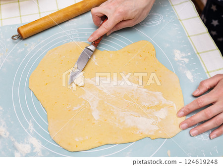 A young Caucasian female baker spreads butter on sweet dough, preparing Easter bread. She uses a knife to evenly distribute the filling. Preparation for a delicious holiday treat 124619248