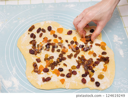 A Caucasian adult female hand adds dried fruits and cranberries to a sweet bread dough, preparing an Easter treat. The dough is spread on a pastry mat, ready for baking 124619249