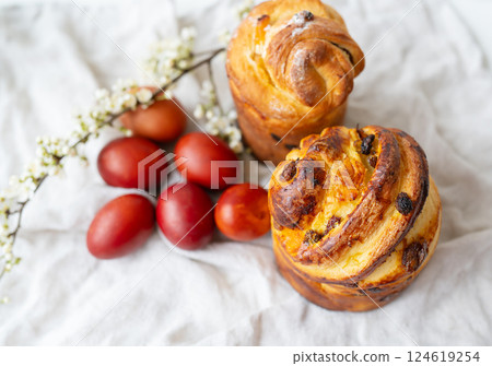 Freshly baked pastries beside red eggs and delicate white flowers, displayed on a textured fabric. 124619254