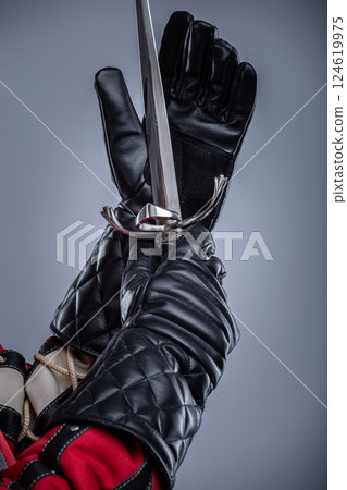 Close-Up of Hands in Black Gloves Holding a Blade. This image provides a detailed view of hands wearing black leather gloves gripping a silver blade with an ornate hilt. Close-Up of Hands in Black Gloves Holding a Blade. This image provides a detailed view of hands wearing black leather gloves gripping a silver blade with an ornate hilt. 124619975