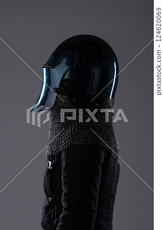 Dark Medieval Helmet with Chainmail Collar. This image shows a dark, almost black, medieval helmet with a perforated visor, paired with a silver chainmail collar and a dark garment underneath. 124620069