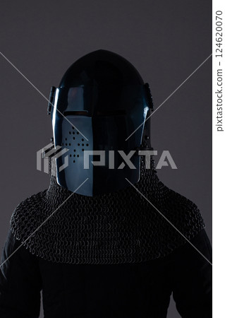 Dark Medieval Helmet with Chainmail Collar. This image shows a dark, almost black, medieval helmet with a perforated visor, paired with a silver chainmail collar and a dark garment underneath. 124620070