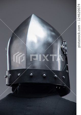 Close-Up of a Medieval Knight's Helmet with Perforated Visor. This image focuses on a shiny metal helmet with numerous small holes in the visor, worn by a person in a dark quilted garment. Close-Up of a Medieval Knight's Helmet with Perforated Visor. This image focuses on a shiny metal helmet with numerous small holes in the visor, worn by a person in a dark quilted garment. 124620074