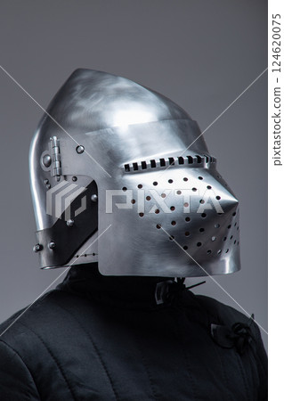 Close-Up of a Medieval Knight's Helmet with Perforated Visor. This image focuses on a shiny metal helmet with numerous small holes in the visor, worn by a person in a dark quilted garment. 124620075