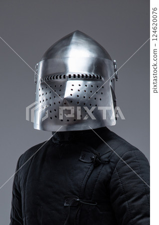 Close-Up of a Medieval Knight's Helmet with Perforated Visor. This image focuses on a shiny metal helmet with numerous small holes in the visor, worn by a person in a dark quilted garment. 124620076