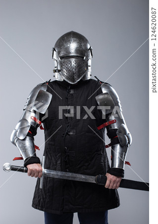 Knight in Full Plate Armor Holding a Sword. This image shows a person fully clad in shiny silver plate armor, including a helmet with a perforated visor, gauntlets, and articulated leg and arm pieces, 124620087