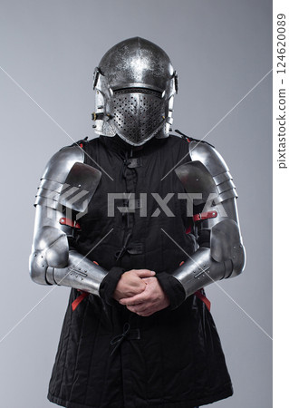 Knight in Full Plate Armor. This image shows a person fully clad in shiny silver plate armor, including a helmet with a perforated visor, gauntlets, and articulated leg and arm pieces, over a dark 124620089