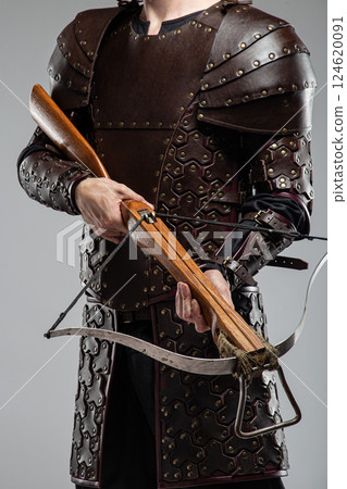Warrior in Full Leather Armor Holding a Crossbow. This image shows a person wearing a complete set of brown leather armor with studded details, holding a wooden crossbow with a metal prod. 124620091