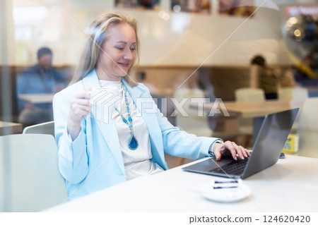 Business woman drinks coffee and works on laptop while sitting at table in cafe. View through window 124620420