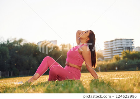 In pink clothes. Sitting on the ground. Beautiful woman in sportive clothes doing fitness exercises outdoors on the field 124622986