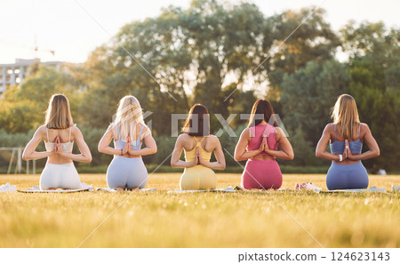 Rear view. Sitting in lotus pose with hands on the back. Group of women have fitness outdoors on the field together 124623143