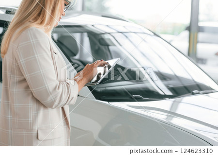 With notepad in hands. Woman in white formal clothes is in the car dealership 124623310
