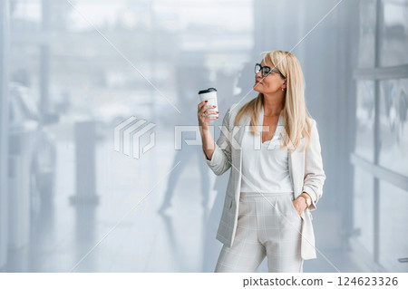 Against grey background with drink in hand. Woman in white formal clothes is standing indoors 124623326