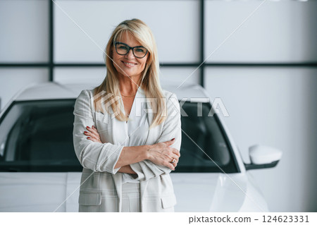 Standing with arms crossed. Woman in white formal clothes is in the car dealership 124623331