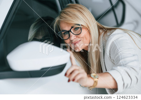 Leaning on the automobile. Woman in white formal clothes is in the car dealership Leaning on the automobile. Woman in white formal clothes is in the car dealership 124623334
