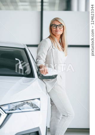 Leaning on the automobile. Woman in white formal clothes is in the car dealership Leaning on the automobile. Woman in white formal clothes is in the car dealership 124623344