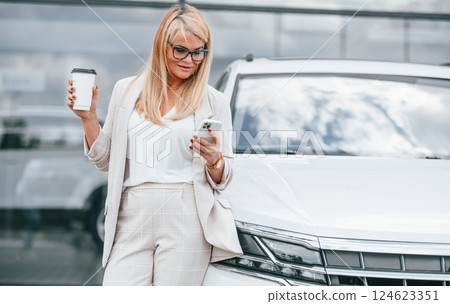 Outdoors with cup of drink. Woman in white formal clothes is in the car dealership Outdoors with cup of drink. Woman in white formal clothes is in the car dealership 124623351