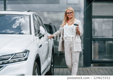 Outdoors with cup of drink. Woman in white formal clothes is in the car dealership Outdoors with cup of drink. Woman in white formal clothes is in the car dealership 124623352