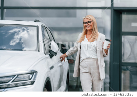 Outdoors with cup of drink. Woman in white formal clothes is in the car dealership 124623353
