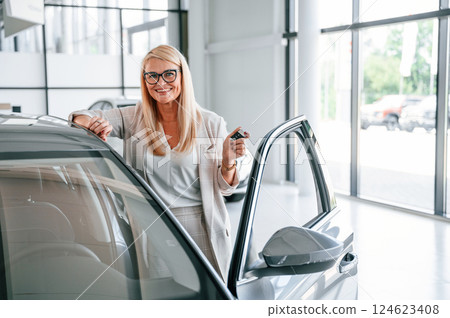 Happy owner of new automobile with keys in hand. Woman in white formal clothes is in the car dealership 124623408