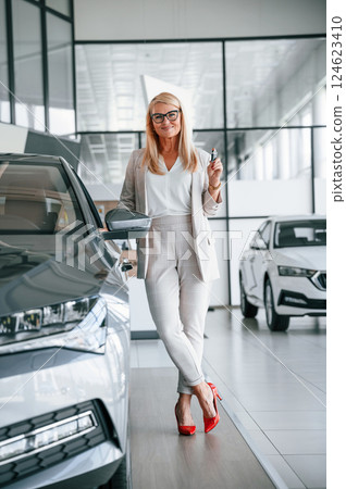 Happy owner of new automobile with keys in hand. Woman in white formal clothes is in the car dealership 124623410