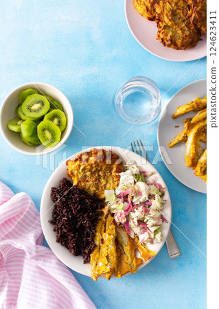 A healthy breakfast featuring brown rice with baked white fish, zucchini-carrot pancakes, Chinese cabbage salad with radish, and a refreshing kiwi dessert for a balanced, nutritious start to the day 124623411