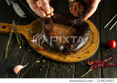 A pair of hands adds seasoning to a dark sausage resting on a wooden board. Surrounding ingredients include garlic, herbs, and chili, enhancing the culinary atmosphere 124624083