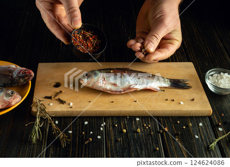 Hands are seen preparing a fresh fish on a wooden cutting board. Spices and herbs are being sprinkled on top while other ingredients rest nearby. The atmosphere is cozy and inviting 124624086