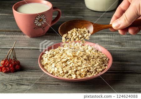 A hand holds a wooden spoon, pouring oatmeal into a pink plate, with a cup of milk nearby. The kitchen has a warm ambiance, showcasing simple, wholesome breakfast preparation A hand holds a wooden spoon, pouring oatmeal into a pink plate, with a cup of milk nearby. The kitchen has a warm ambiance, showcasing simple, wholesome breakfast preparation 124624088