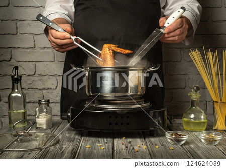 In a modern kitchen, a chef skillfully uses a fork and knife to handle the chicken legs cooking in a pan. The warm atmosphere is illuminated by the stove In a modern kitchen, a chef skillfully uses a fork and knife to handle the chicken legs cooking in a pan. The warm atmosphere is illuminated by the stove 124624089