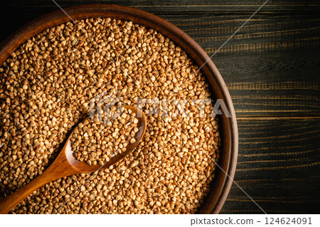 A wooden bowl filled with golden buckwheat grains sits on a dark wooden surface. A wooden spoon rests on the edge, highlighting the natural beauty and texture of the seeds. 124624091