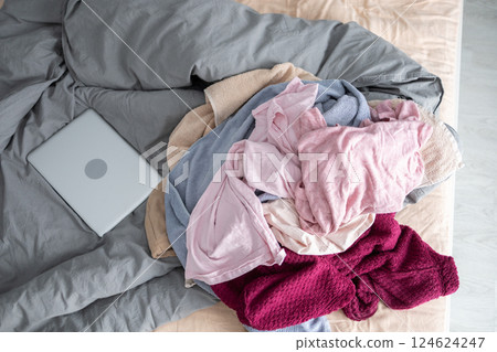 Messy pile of clothes and laptop on unmade bed in bedroom. Household clutter and everyday disorganization in a lived-in home. 124624247