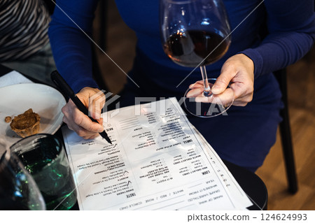 Close-up of a woman filling out a tasting sheet while holding a glass of red wine during a workshop. Wine knowledge, consumer empowerment, wine literacy, educated choices Close-up of a woman filling out a tasting sheet while holding a glass of red wine during a workshop. Wine knowledge, consumer empowerment, wine literacy, educated choices 124624993
