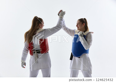Smiling female taekwondo fighters in red and blue gear celebrating with high five after match on white background. 124625432