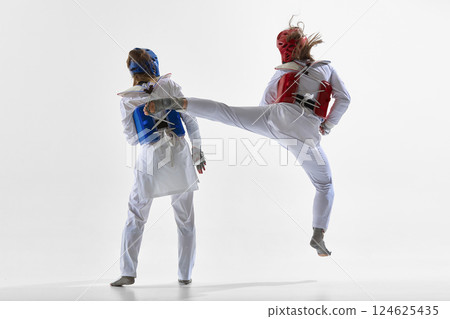 Woman in red gear jumping mid-air to strike blue female opponent with powerful side kick during match on white background. Woman in red gear jumping mid-air to strike blue female opponent with powerful side kick during match on white background. 124625435