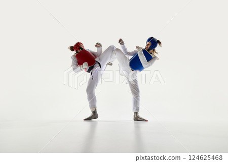 Female taekwondo fighters in red and blue protective gear in motion, fighting with synchronized kicks on white background. 124625468