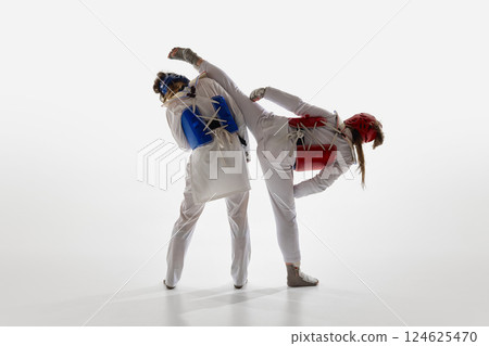 Red taekwondo athlete performing high kick behind blue opponent, two women practicing against white background. 124625470