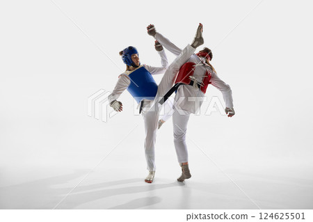 Two young women, taekwondo fighters in blue and red gear in motion, in dynamic move, training against white background. Two young women, taekwondo fighters in blue and red gear in motion, in dynamic move, training against white background. 124625501
