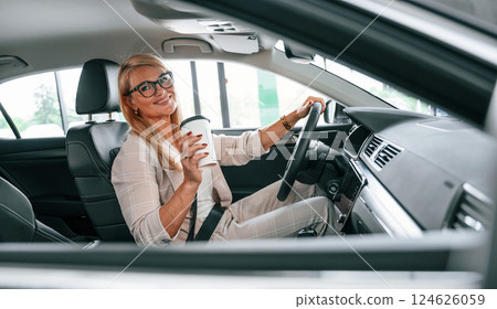 Driving vehicle and holding cup of drink. Woman in white formal clothes is in the car dealership 124626059