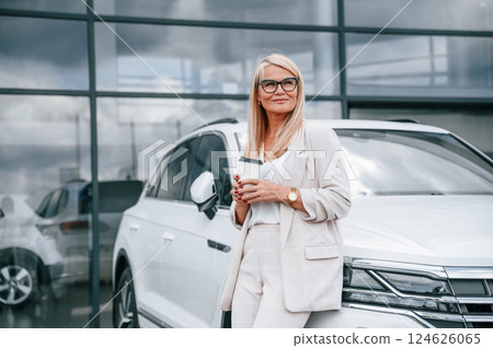 Outside with cup of drink. Woman in white formal clothes is in the car dealership 124626065