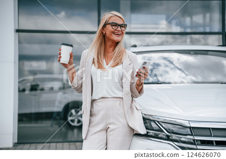 Standing against building with big windows. Woman in white formal clothes is in the car dealership 124626070