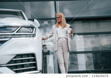 Standing outdoors against building and holding drink. Woman in white formal clothes is in the car dealership 124626074