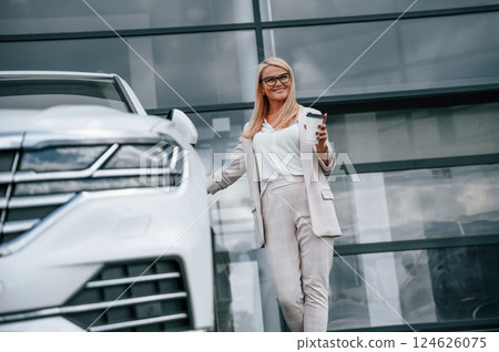 Standing outdoors against building and holding drink. Woman in white formal clothes is in the car dealership 124626075