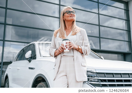 Standing outdoors near new automobile. Woman in white formal clothes is in the car dealership Standing outdoors near new automobile. Woman in white formal clothes is in the car dealership 124626084