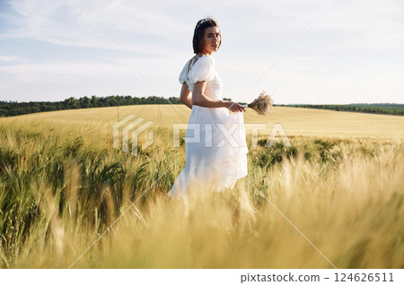 Holding flowers. Beautiful young bride in white dress is on the agricultural field at sunny day 124626511