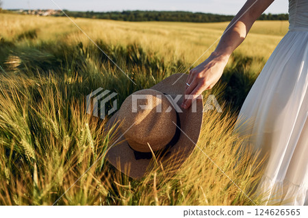 Holding hat. Beautiful young bride in white dress is on the agricultural field at sunny day 124626565