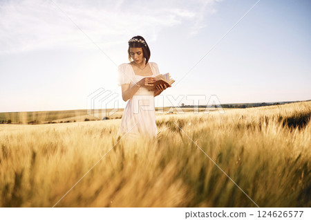 With book in hands. Beautiful young bride in white dress is on the agricultural field at sunny day With book in hands. Beautiful young bride in white dress is on the agricultural field at sunny day 124626577