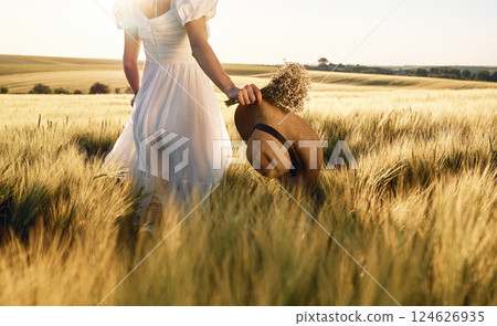 Cropped view. Holding hat in hand. Beautiful young bride in white dress is on the agricultural field at sunny day 124626935